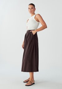 Brauner, hoch taillierter, plissierter Rock mit Taschen; kombiniert mit einem engen, weißen gerippten Tanktop und schwarzen flachen Sandalen. Glatte Textur.