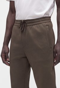 Σκούρο καφέ φόρμα Armani Exchange με κορδόνι στη μέση και πλαϊνές τσέπες, φορεμένη από άτομο με λευκό πουκάμισο.