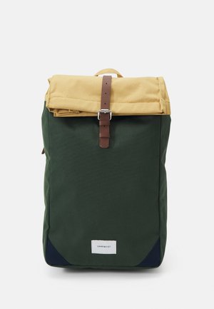 Grüner und beiger Rucksack mit einer oberen Klappe, die durch einen braunen Schnallenriemen gesichert ist, und einem kleinen weißen Logoschild vorne unten.