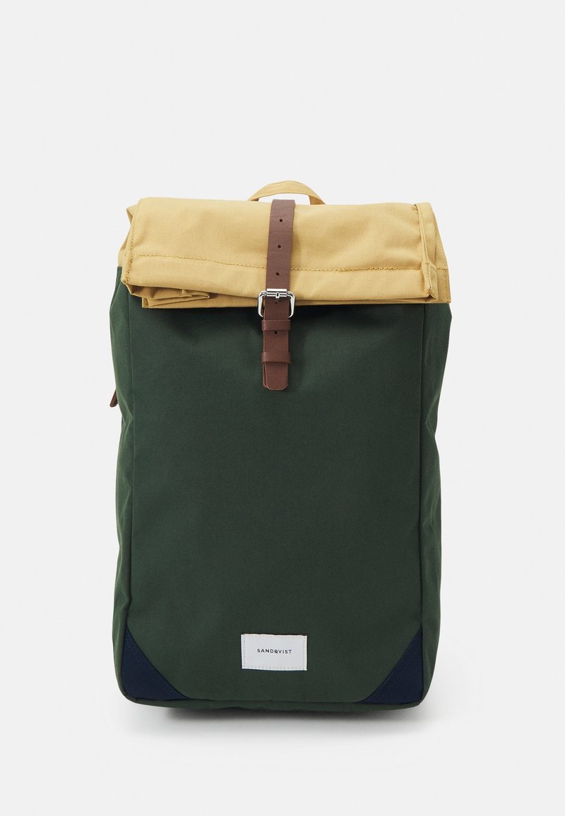 Sac à dos vert et beige avec un rabat supérieur sécurisé par une sangle à boucle marron et un petit écusson blanc avec logo en bas à l'avant.