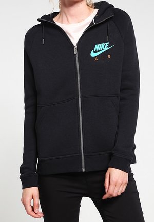 Personne portant un sweat à capuche zippé Nike Air noir avec un logo bleu clair et des poches avant, associée à un pantalon noir.