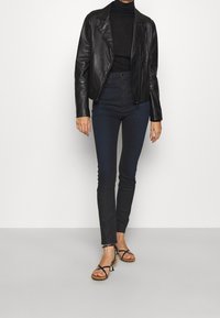 Černá kožená motocyklová bunda přes černý rolák, spárovaná s tmavými úzkými džíny a černými sandály na podpatku. Model stojí ve středu.