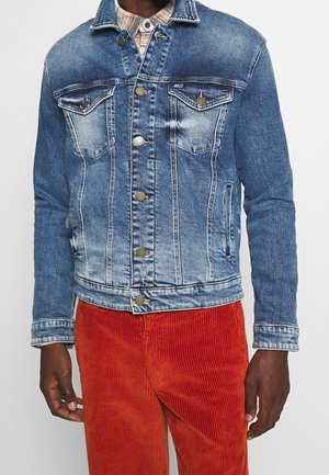 Man draagt een blauwe spijkerjack met koperen knopen over een geruit overhemd en rode ribfluwelen broek.