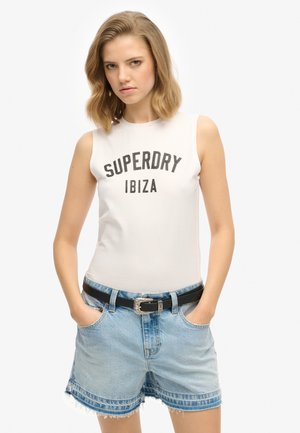 Top sin mangas blanco con estampado en negro que dice "SUPERDRY IBIZA"; combinado con pantalones cortos de mezclilla azul claro y un cinturón negro con hebilla plateada.