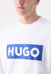 Balta sportinė palaidinė su gumine apykakle, ant kurios yra didelė mėlyna stačiakampio formos grafika su užrašu "HUGO" ryškiomis baltomis raidėmis.