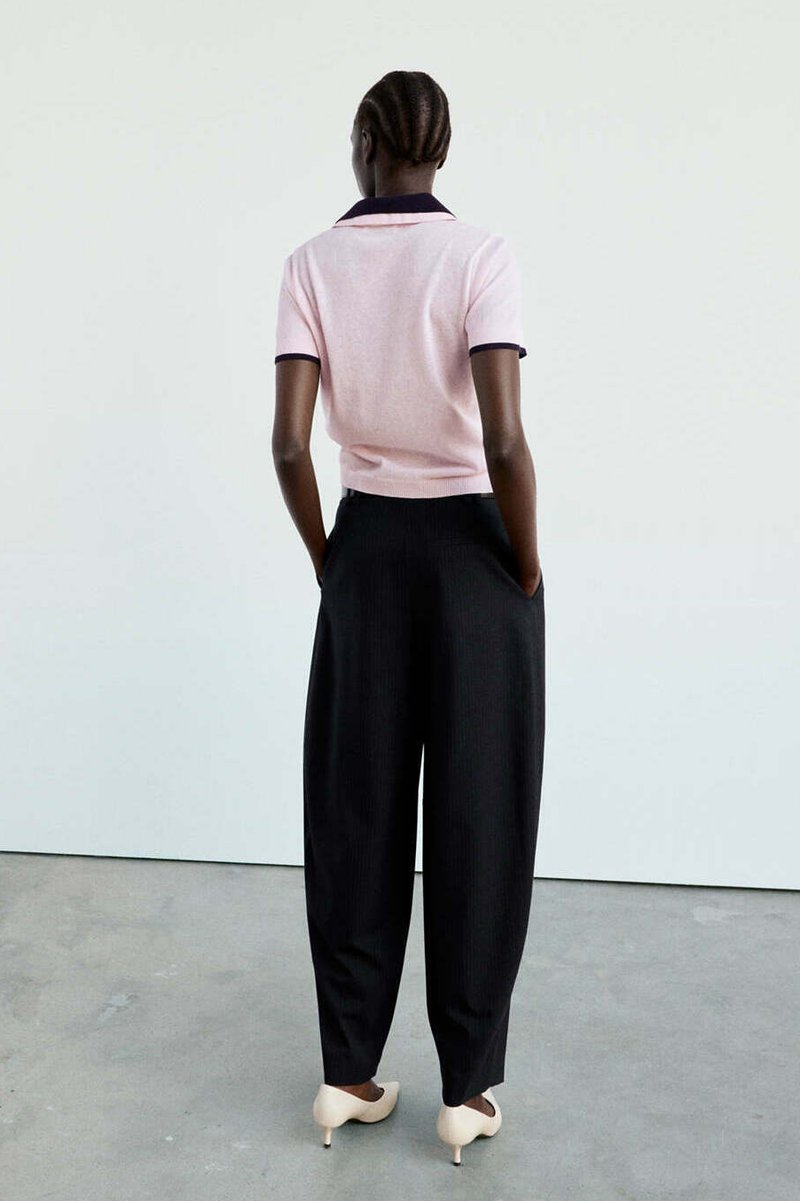 Personne avec de courts cheveux tressés portant une chemise rose à manches courtes, un pantalon noir large et des talons beiges, debout les mains dans les poches.