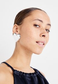 Pendientes de plata en forma de corazón, con una superficie lisa y reflectante, llevados por una persona de tez media y piel con pecas.