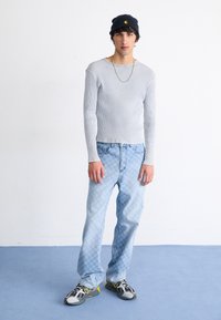 Maglia a maniche lunghe ribbed grigia chiara, jeans a fantasia blu chiaro, sneakers grigie e gialle, berretto nero con logo, in piedi su uno sfondo neutro.