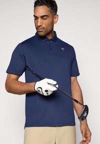 Ναυτικό μπλουζάκι πόλο με γιακά και τρία κουμπιά, συνδυασμένο με λευκό γάντι γκολφ με μαύρες λεπτομέρειες, κρατώντας ένα μπαστούνι γκολφ.