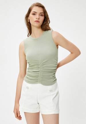 Hellgrünes ärmelloses Oberteil mit gerafften Details an den Seiten, aus weichem Material gefertigt. Kombiniert mit weißen Shorts mit silbernen Akzenten.