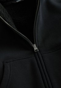 Schwarze Fleecejacke mit Reißverschluss, der teilweise geöffnet ist, sodass das weiche, strukturierte Innenfutter und die Nähte der Fronttasche sichtbar sind.