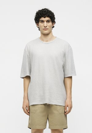 Νέος άνδρας με κοντά σγουρά σκούρα μαλλιά, φορώντας γκρι και λευκό ριγέ φαρδιά μπλούζα και χακί σορτς, στέκεται μπροστά σε απλό φόντο.