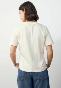 T-shirt à manches courtes de couleur crème en coton doux. Présente une coupe droite et une couture centrale dans le dos. Jeans visibles en bas.