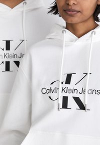 Bijela dukserica s velikim crnim grafičkim logotipom "Calvin Klein Jeans" i drugim stiliziranim slovima. Ima kapuljaču s vezicom i kengur džep.