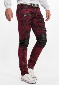 Jeans bordeaux déchirés avec plusieurs zippers, clous et accents en cuir noir. Présente une coupe slim et un tissu texturé.