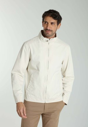 Homme portant une veste blanche zippée avec col montant et pantalon beige, debout devant un fond clair uni.
