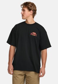Svart oversized t-shirt i bomull med en röd logotyp på vänster bröst. Kort ärm och rund hals. Slät textur.