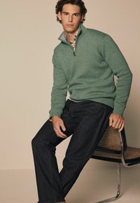 Grüner Wollpullover mit kurzem Reißverschluss, strukturierte Oberfläche, über einem Hemd mit Kragen getragen. Dunkelblaue Jeans im Straight-Leg-Schnitt, glatte Oberfläche, minimale Nähte.