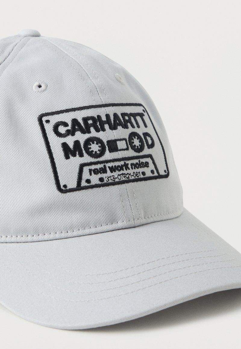Gorra de béisbol gris claro con un diseño bordado en negro de un cassette que dice "CARHARTT MOOD raw work noise" en el panel frontal.