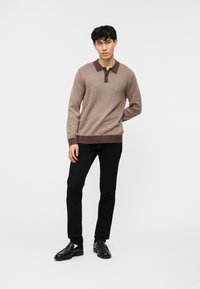 Νέος άνδρας που στέκεται με καφέ μακρυμάνικη μπλούζα πόλο, μαύρο παντελόνι και μαύρα επίσημα παπούτσια, πάνω σε απλό λευκό φόντο.