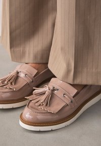 Mocassins en cuir verni rose pâle avec des pampilles, dotés d'une semelle blanche et de ferrures argentées, associés à un pantalon rayé beige.