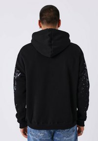 Felpa nera con cappuccio grande, caratterizzata da un design a macchie grigie su entrambe le maniche. Realizzata in materiale morbido con polsini e orlo a coste.