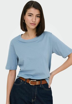Young woman with dark bob hair, wearing gold hoop earrings, a light blue short-sleeve t-shirt, dark jeans, and a brown belt, posing with hand on hip.
