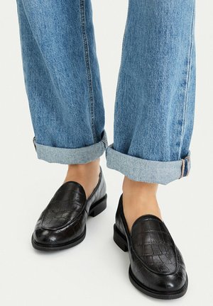 Person iført op-rullede lyseblå jeans og sorte teksturerede læderloafers på en lys baggrund.