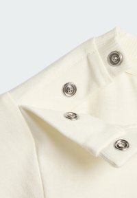 Detail zblízka bielej látky so tromi striebornými kovovými cvokmi, z ktorých jeden je rozopnutý, pričom sú vidieť dierky na cvoky a detaily stehu.