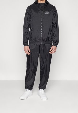 Mężczyzna ubrany w czarną bluzę z kapturem na zamek błyskawiczny i pasujące spodnie oraz białe trampki, stoi na jednolitym jasnym tle.