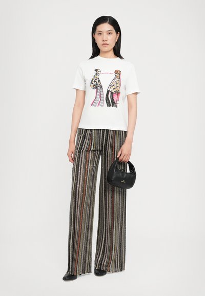 T-shirt blanc à manches courtes avec un design graphique, associé à un pantalon large rayé en noir, or et couleurs. Le mannequin tient un petit sac noir.