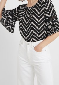 Schwarze Bluse mit Chevron-Muster und weißen Blumenmotiven, mit Rüschenärmeln, kombiniert mit hoch taillierten weißen Jeans.