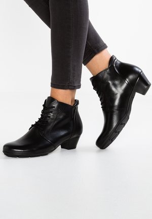 Bottes noires en cuir montant à la cheville avec talons bas et lacets, portées avec un pantalon noir ajusté sur fond blanc.