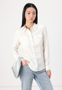 White, long-sleeve button-up shirt with two chest pockets, paired with light blue jeans and holding a silver clutch. Smooth fabric texture.