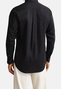 Personne portant une chemise noire ajustée à manches longues et un pantalon de couleur claire, vue de dos, avec les mains détendues sur les côtés.