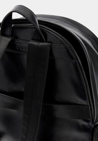 Sac à dos en cuir noir avec finition texturée, doté d'une poignée supérieure, de bretelles en nylon, d'une fermeture éclair en métal et de détails sur le compartiment intérieur.