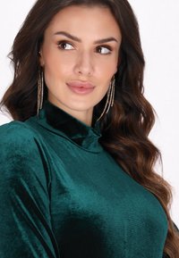 Top din catifea de culoare verde smarald bogat, cu guler înalt. Prezintă o textură netedă și un luciu subtil. Păr lung, cercei cu rhinestone care atârnă vizibili.