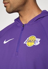 Lila Nike-Hoodie mit einem Reißverschlusskragen, versehen mit einem Los Angeles Lakers-Logo in Gelb und Weiß. Texturiertes Material sichtbar.