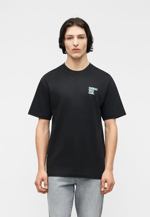 Ung mand med mellem-langt mørkt hår iført en sort T-shirt med teksten "TUESDAY TACO CLUB." øverst til venstre og lyseblåt jeans.