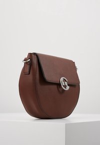 Brązowa okrągła torebka o gładkiej fakturze, ze srebrnymi okuciami i zapięciem na klapę. Na przodzie widoczny jest wyraźny detal z logo.