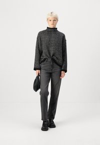 Gestreifter Rollkragenpullover in Schwarz-Weiß mit Chevron-Muster, kombiniert mit dunkelgrauen geraden Jeans und schwarzen Stiefeletten. Hält eine kleine schwarze Tasche.