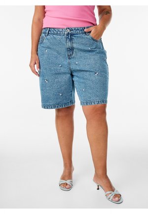 Femme portant un short en jean bleu clair avec de petites perles, un haut rose et des talons ouverts gris clair, debout avec une main dans la poche.