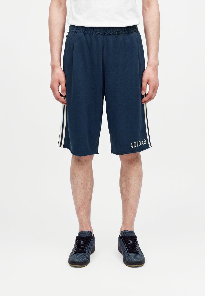 Άτομο που φοράει σκούρα μπλε σορτς Adidas με λευκές πλαϊνές ρίγες, στέκεται με χαλαρά χέρια, συνδυασμένα με μπλε αθλητικά παπούτσια και λευκό t-shirt.