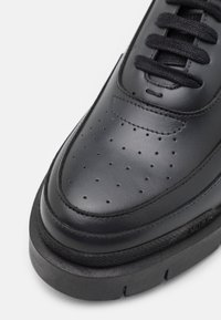 Baskets en cuir noir avec embout perforé, lacets noirs, semelle noire épaisse et coutures détaillées sur fond blanc.
