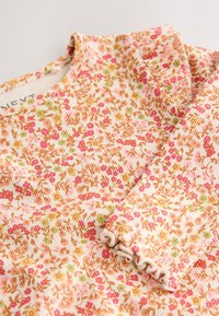 Mäkký vrúbkovaný top s dlhými rukávmi s drobnými kvetinovými potlačami v oranžových, ružových, zelených a žltých tónoch na krémovom pozadí.