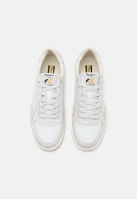 Baskets blanches en cuir avec une texture lisse, orteil perforé et surpiqûres beiges. Elles possèdent des lacets plats et un logo sur la languette.