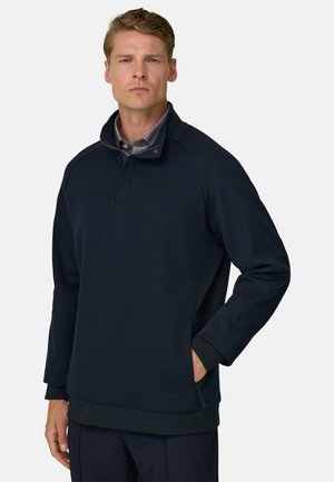 Mand iført en mørkeblå pullover med lynlåslukning ved kraven og en stribet skjorte under, stående med den ene hånd i lommen.