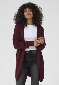 Cardigan en tricot bordeaux avec un devant ouvert, motif texturé, manches longues et poignets côtelés, associé à un haut blanc et un jean noir.