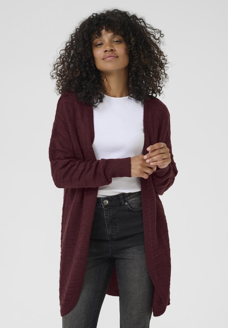 Cardigan en tricot bordeaux avec un devant ouvert, motif texturé, manches longues et poignets côtelés, associé à un haut blanc et un jean noir.