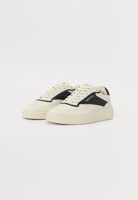 Baskets de couleur crème avec des accents noirs, présentant un haut texturé, une semelle épaisse et un design classique à lacets.